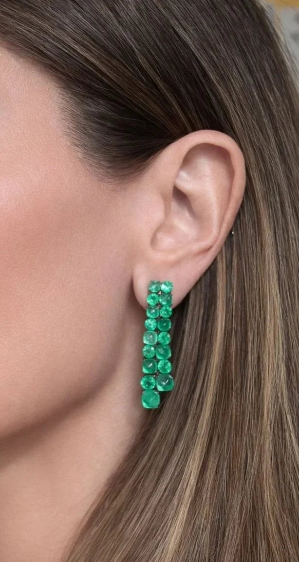 A close-up of a model's ear wearing a vertical drop earring made of multiple round green gemstones.