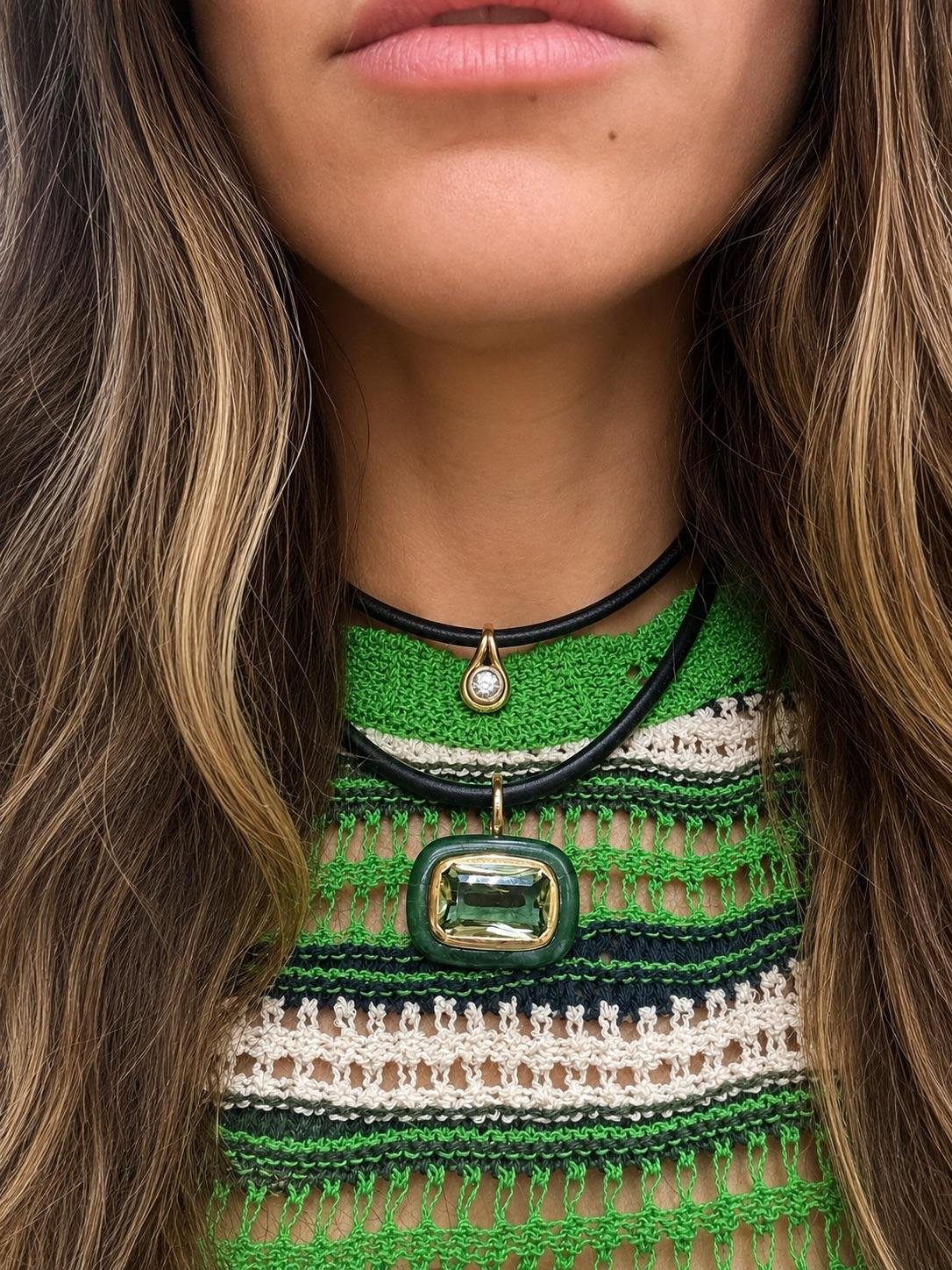 Close-up of a woman wearing a green and cream striped knit top, featuring a layered necklace with a black cord, gold-toned accents, and a prominent green rectangular pendant.