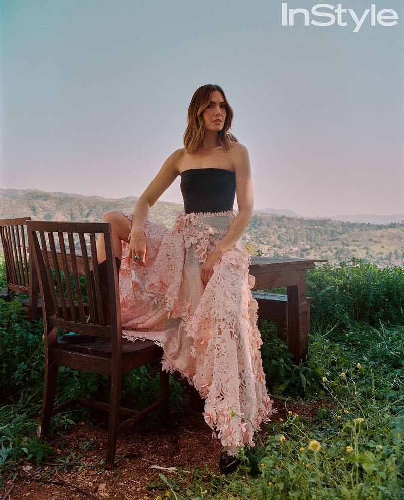 A woman sitting outdoors on a wooden chair, wearing a black strapless top and a flowing pink skirt with floral lace details.