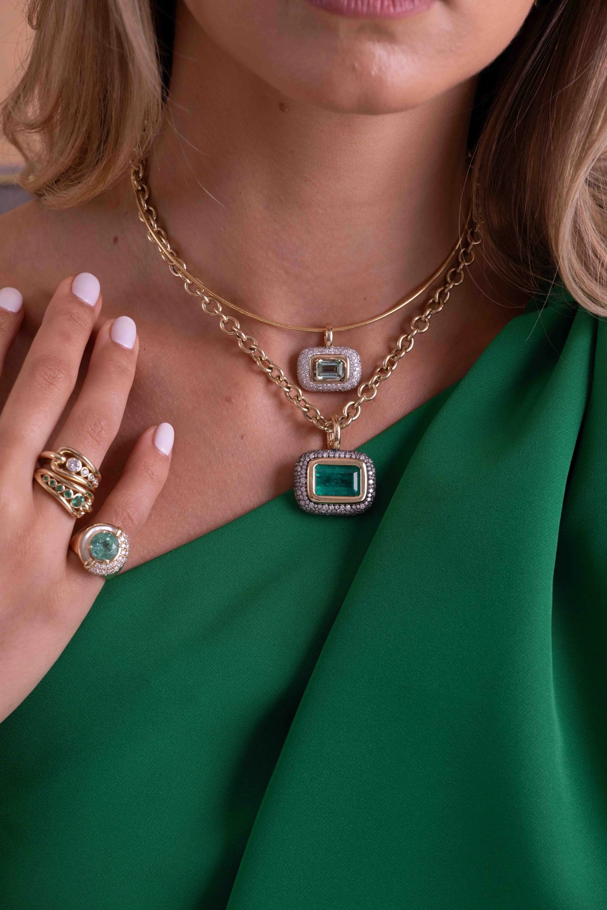 A close-up lifestyle shot of a model wearing a gold double-strand necklace with a rectangular emerald-cut green gemstone pendant, along with matching gold rings and a green dress.