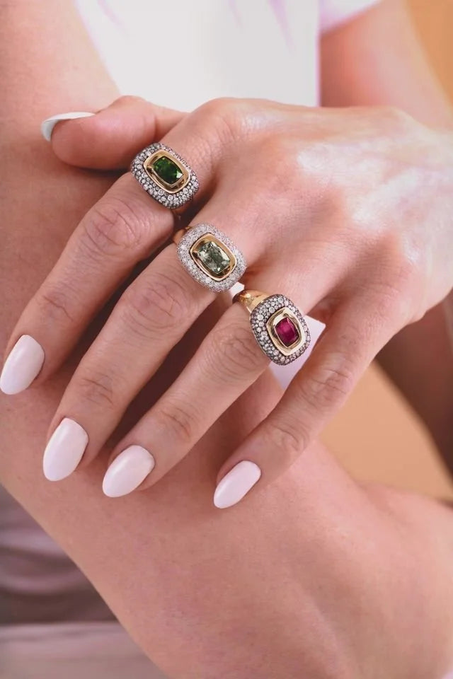 Three gold rings with green and red gemstones and diamond halos worn on a person's hand with pale pink manicured nails.