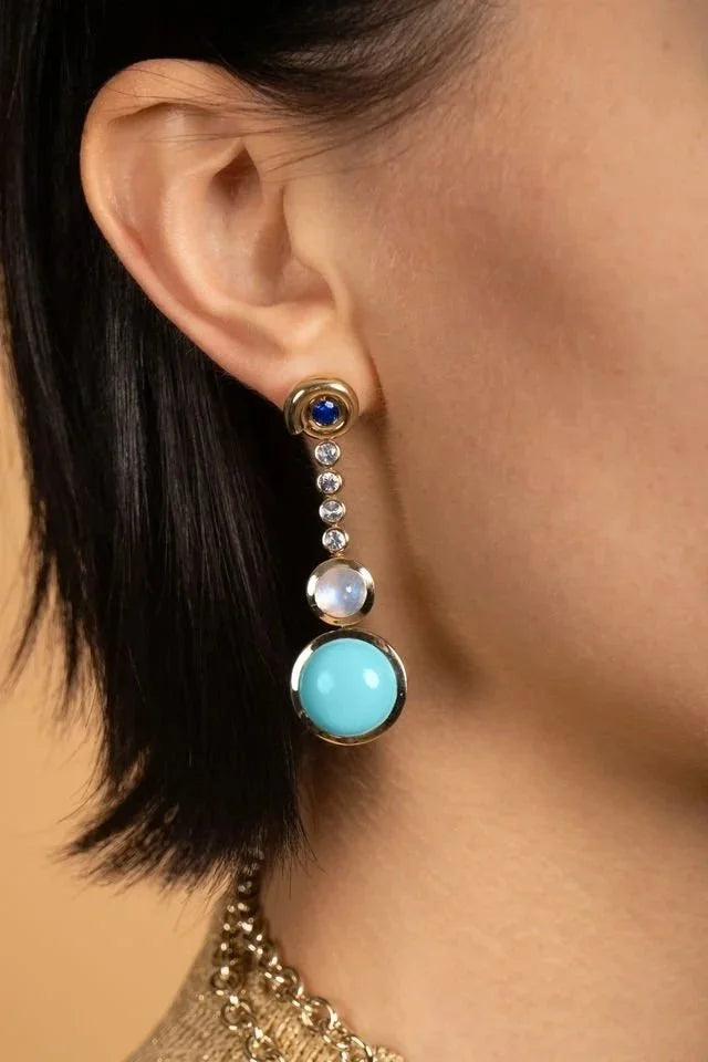 A close-up of a model's ear wearing a dangling earring with a gold-tone setting, a dark blue round stone, clear spacer stones, a white pearl-like bead, and a large light blue oval stone.