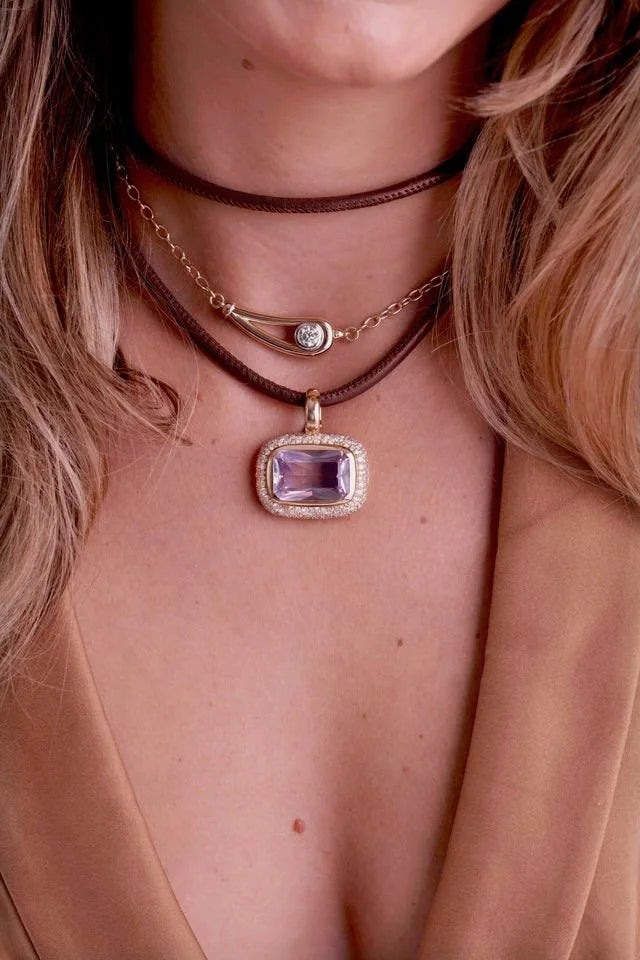 A close-up of a model wearing a multi-layered necklace set. The set includes a dark brown leather choker, a gold chain with a small silver-toned curved charm, and a gold pendant featuring a rectangular purple amethyst stone surrounded by a clear crystal halo.