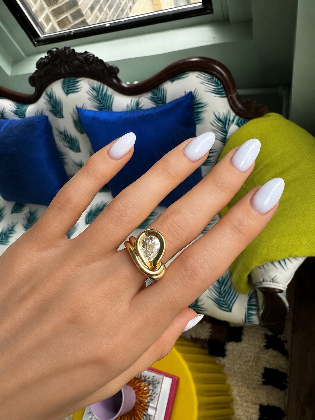 A gold heart-shaped ring with a clear gemstone is worn on a hand with white manicured nails, set against a cozy indoor background with blue and green pillows.