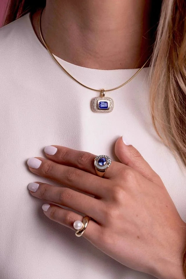 A close-up lifestyle shot of a woman wearing a gold necklace with a rectangular blue gemstone pendant, along with two gold rings on her fingers, set against a light pink top.