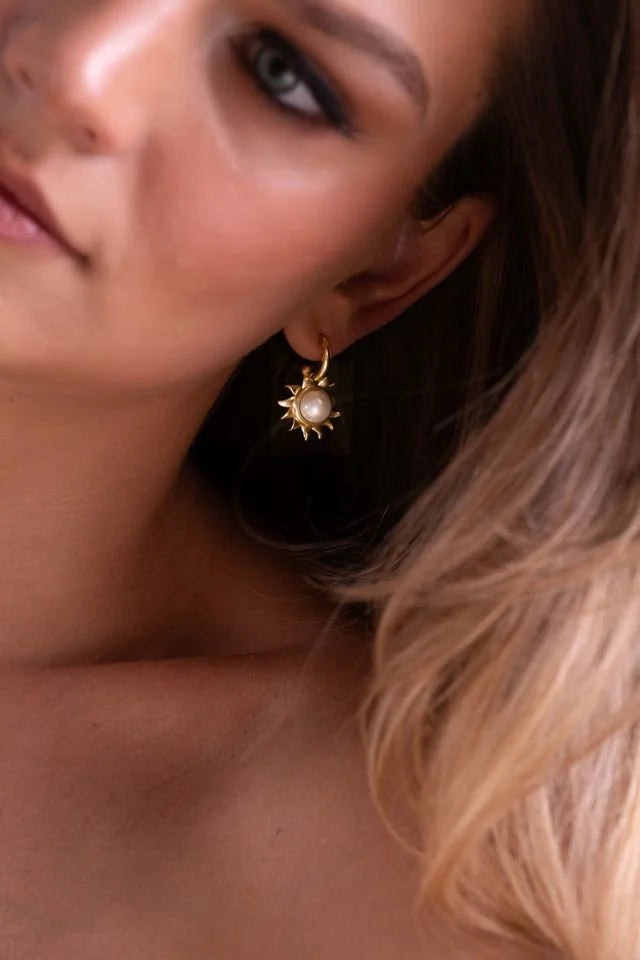 A close-up lifestyle shot of a model wearing a gold sunburst dangle earring with a central white pearl.