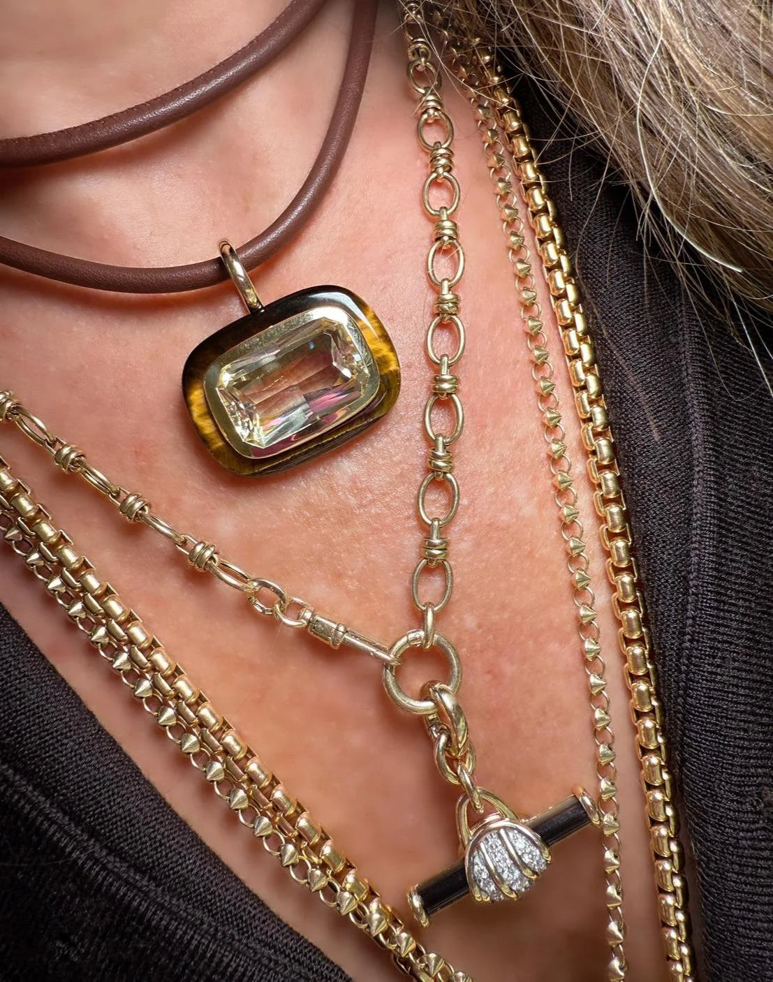 A close-up of a layered necklace set worn on a model's neck, featuring a gold-toned rectangular bezel pendant with a clear faceted gemstone, a brown leather cord layer, and multiple gold chain styles.