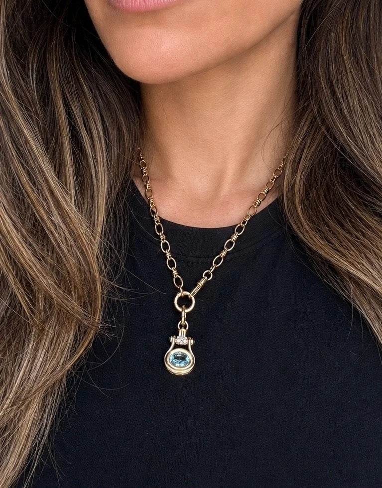 A close-up of a model wearing a gold chain necklace with a small round pendant featuring a light blue gemstone, styled with a black top.