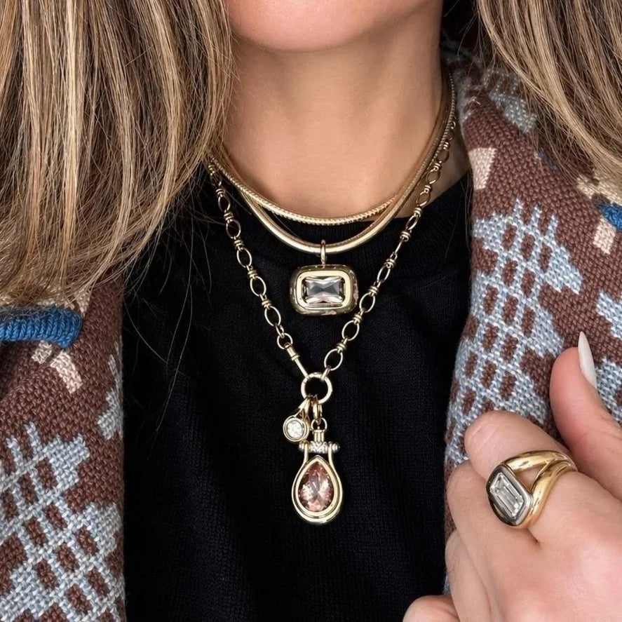 A close-up of a person wearing a layered gold necklace set with a teardrop pendant and a small charm, styled with a patterned brown cardigan and a gold ring.