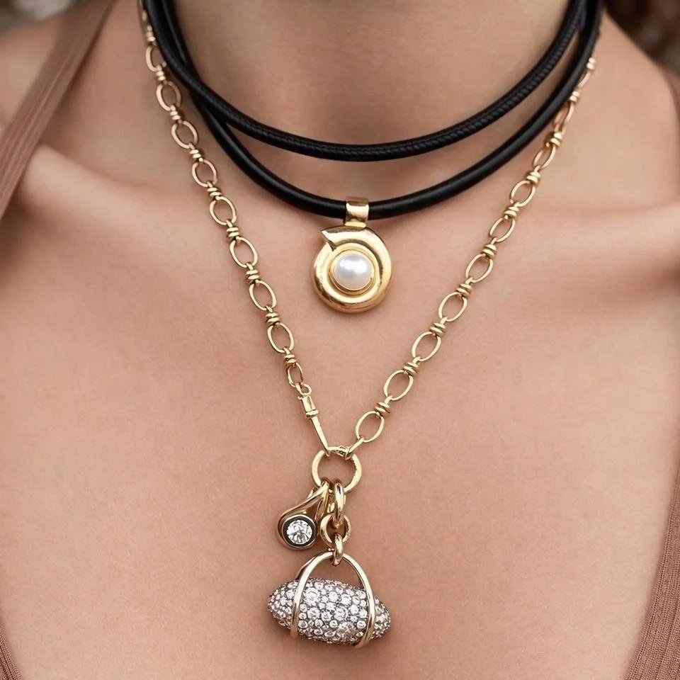 A close-up of a layered necklace worn on a model's neck, featuring a black double-strand cord necklace with a gold pendant and a gold chain necklace with a gold and silver purse-shaped charm.