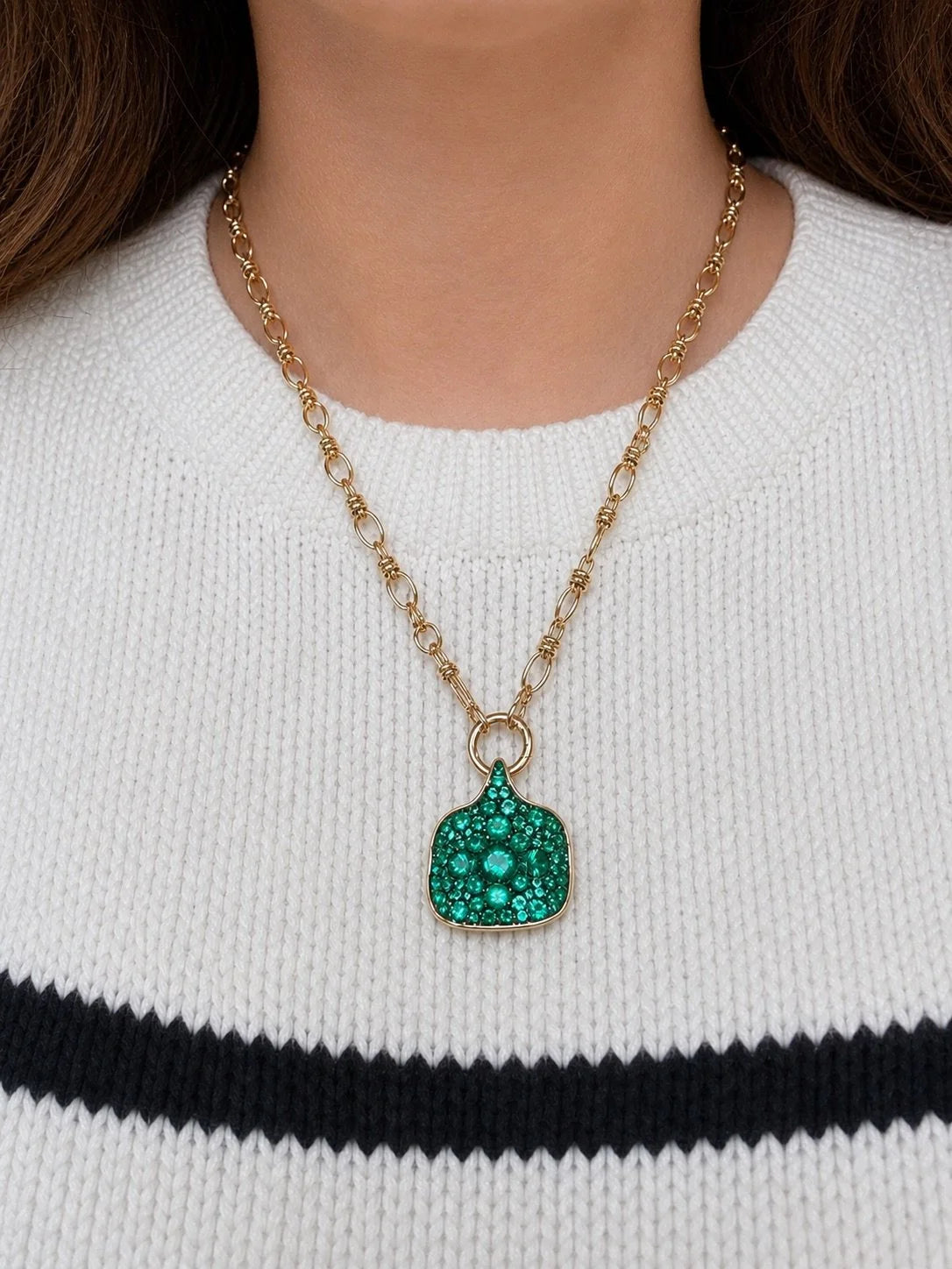 A gold chain necklace with a green textured pendant, worn by a model in a white knit sweater with a black stripe.