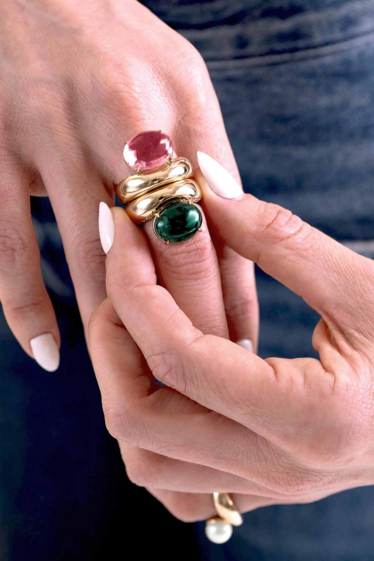 A close-up of a person's hands wearing a gold ring set with a pink cabochon stone, a green emerald-cut stone, and a pearl accent, against a dark denim background.
