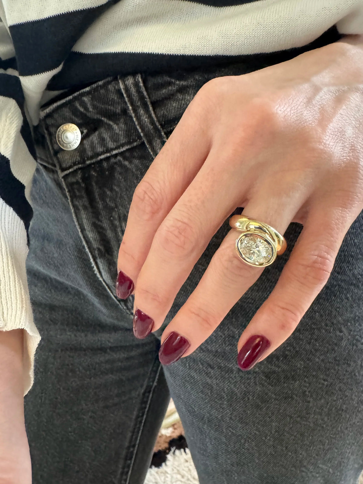A close-up of a person's hand wearing a gold ring with a diamond, styled with dark gray jeans and a black-and-white striped sweater.