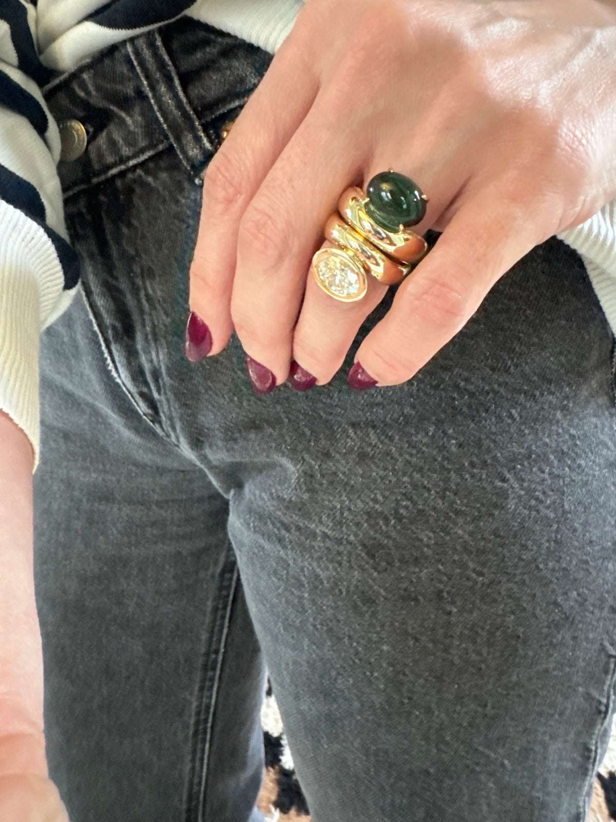 A close-up of a person's hand wearing a stack of three gold rings, including a green oval stone ring and a round clear gemstone ring, paired with dark gray jeans.