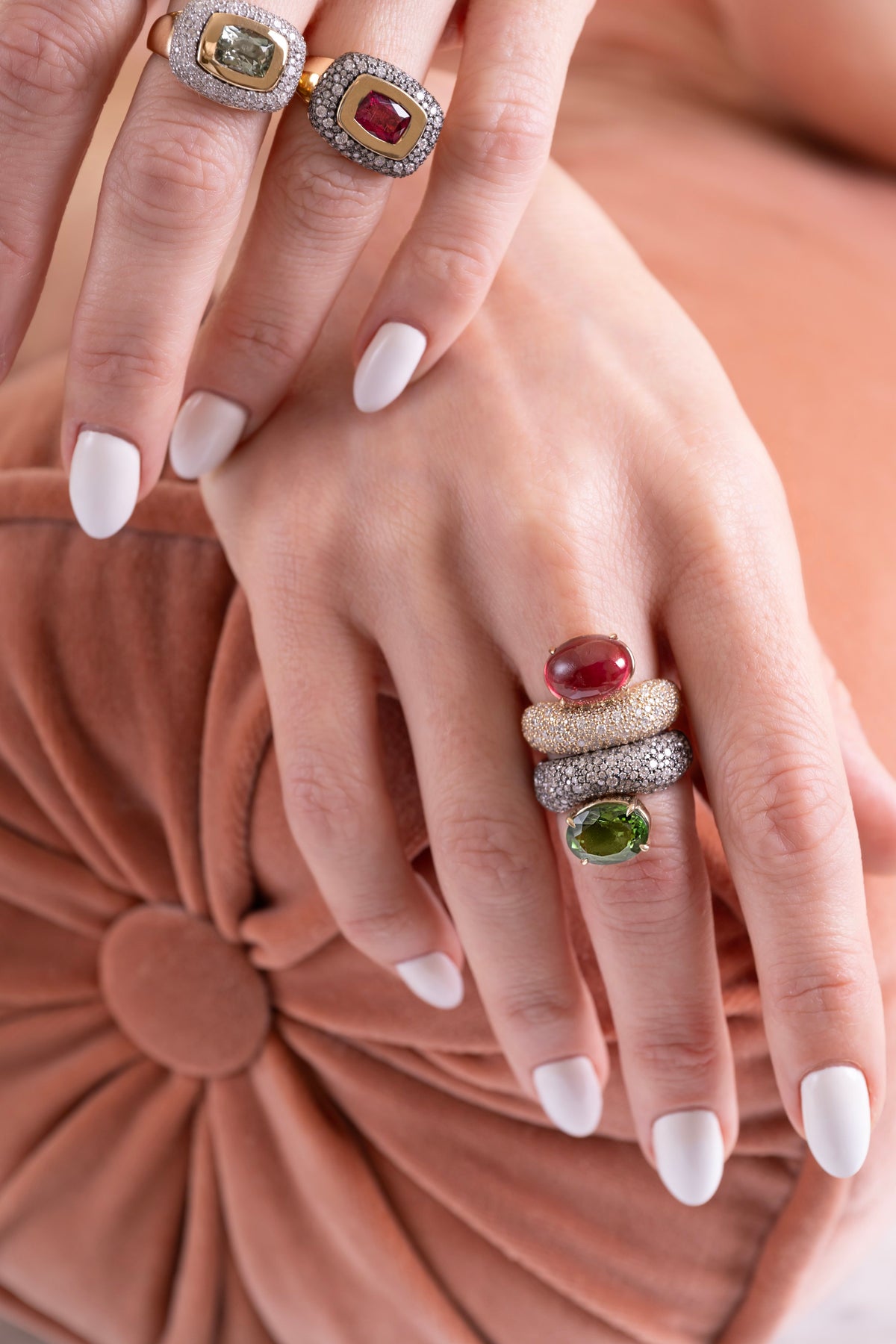 A close-up of a person's hands wearing multiple rings. The hands have white manicured nails and are resting on a dusty rose velvet surface.