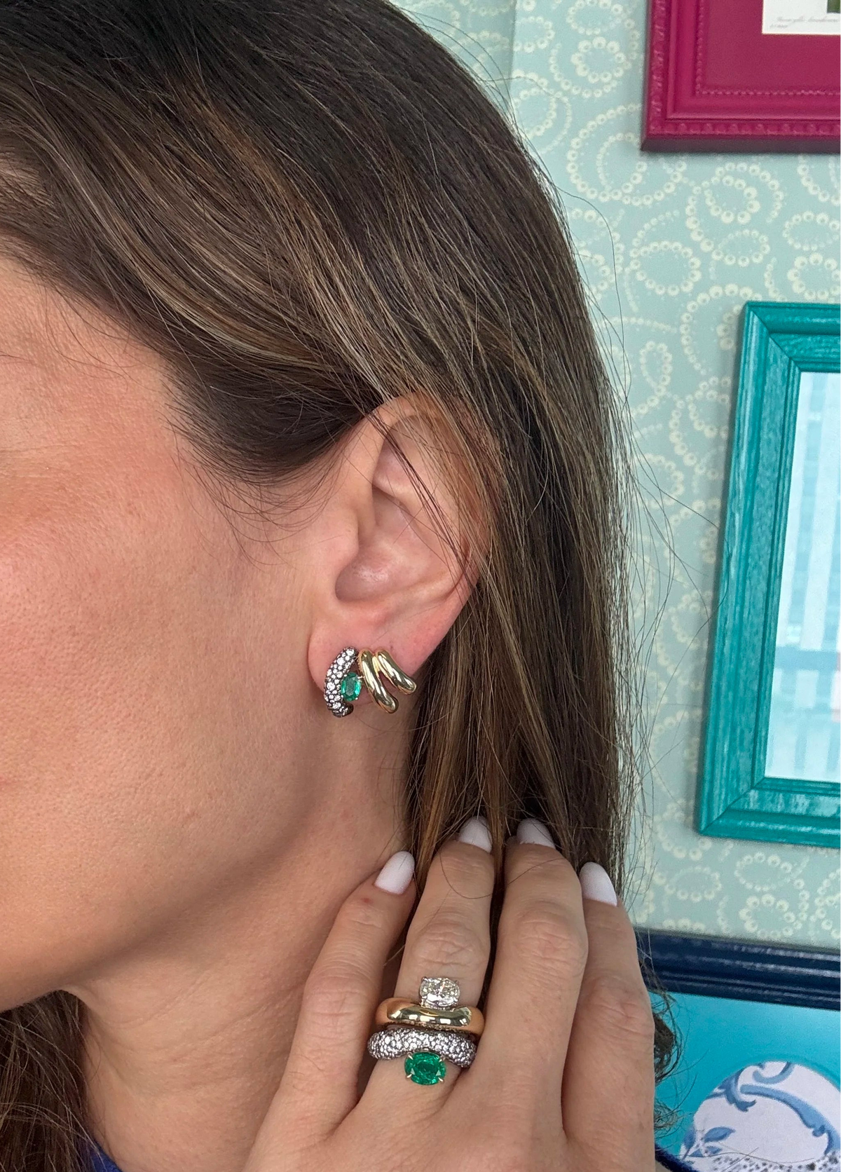 A close-up of a woman wearing a gold ear climber earrings with green gemstone accents, along with stacked rings on her finger, set against a patterned teal wall.