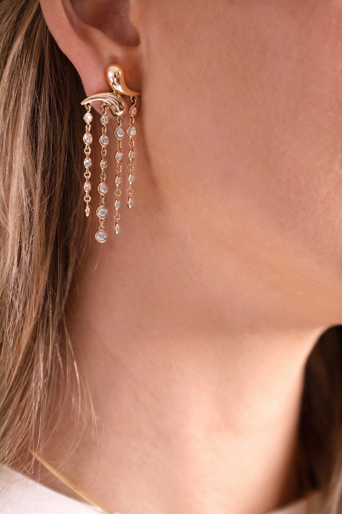 A close-up of a model's ear wearing a gold dangle earring with multiple chains of clear diamonds.
