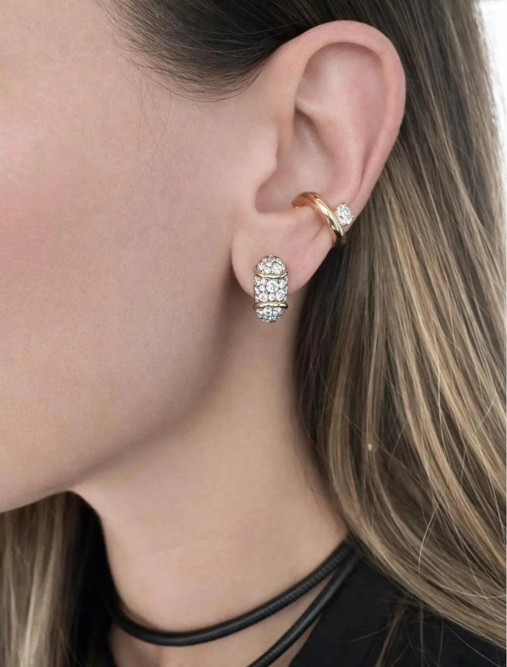 A close-up of a model's ear showcasing a stack of three gold ear cuffs: a small stud earring with clear crystals, a gold cartilage hoop with a clear stone, and a larger gold cuff encrusted with clear crystals.