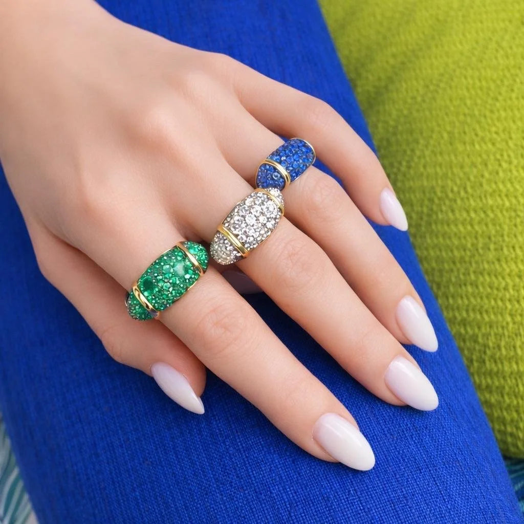 Three gold rings with colorful gemstone inlays are shown on a person's hand. One ring features a vibrant green inlay, one a deep blue inlay, and one is paved with clear stones.